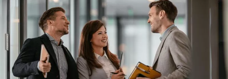 Three business professionals conversing and smiling in a modern office hallway.